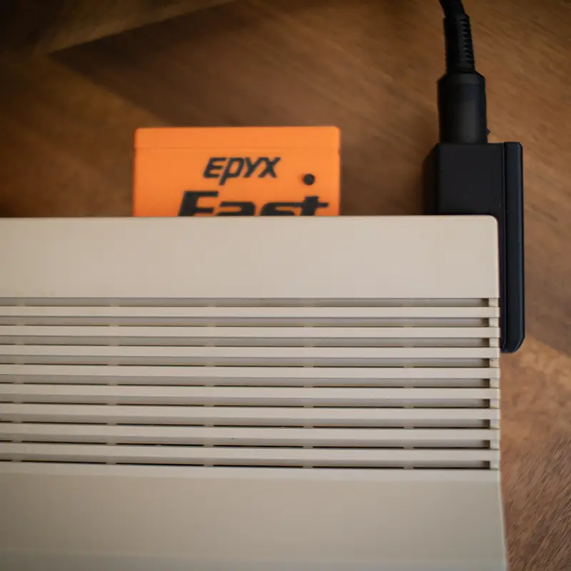 A top down view of a 64C with an inserted Epyx FastLoad cartridge and a right-angle power adapter.